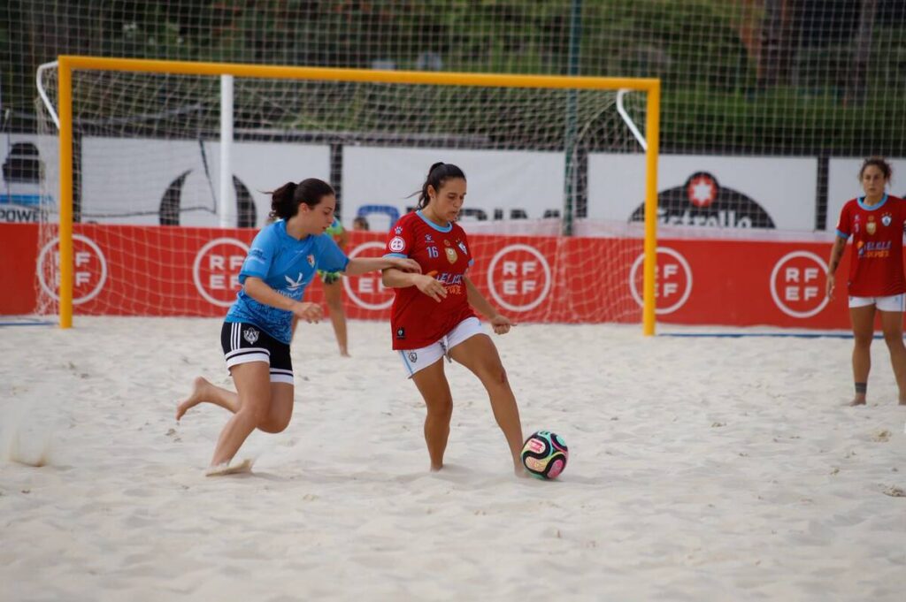 Two female beach soccer players chase the ball near a yellow goal frame on a sandy field with red banners in the background.