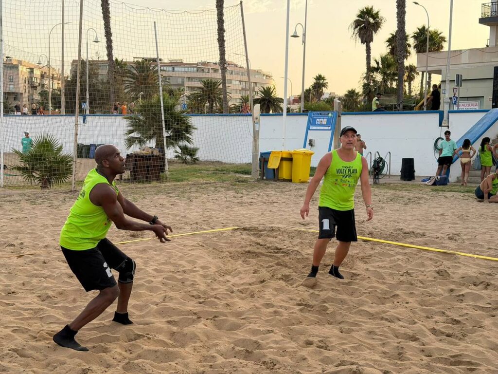 Two men in neon green shirts play beach volleyball on a sandy court with a net in the background.