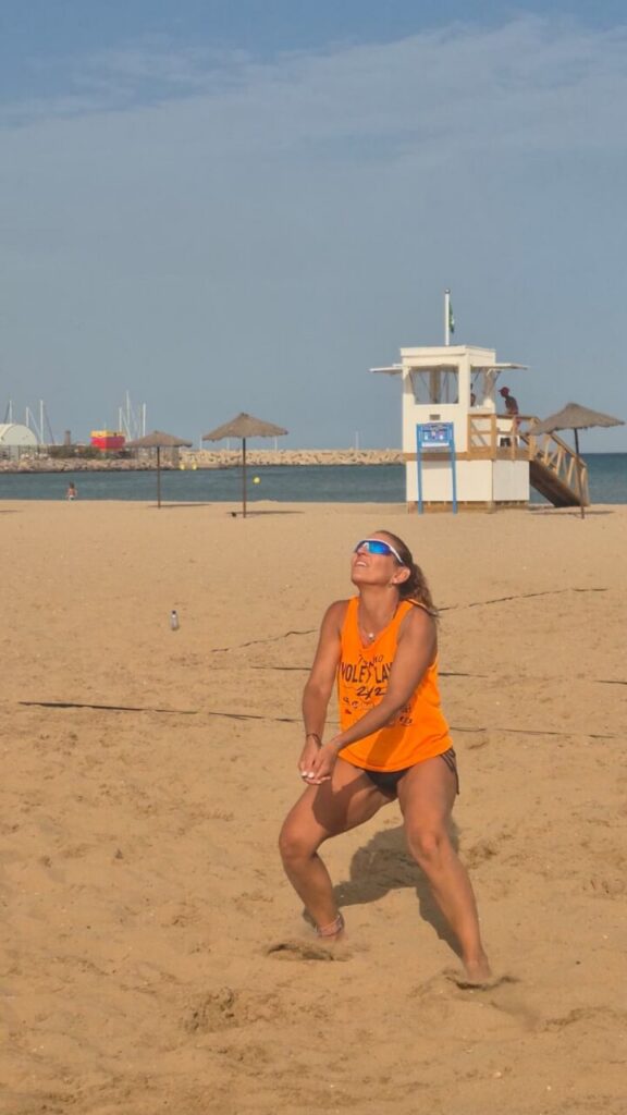 Woman in an orange tank top and sunglasses crouches on a sandy beach, ready to volley a volleyball with the ocean and lifeguard tower in the background.