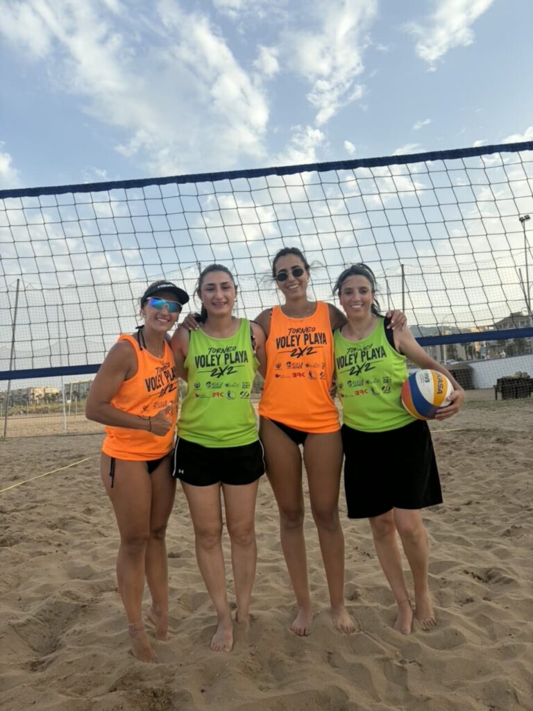 Four women in bright neon jerseys stand together on a sandy beach volleyball court, smiling in front of a net; one holds a volleyball on the right.
