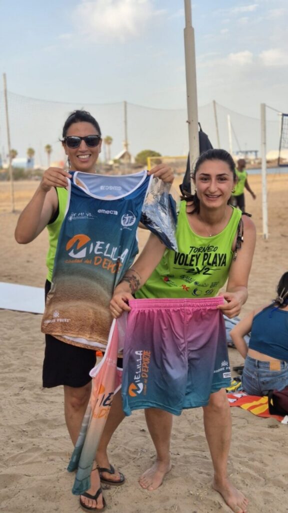 Two smiling women on a sandy beach volleyball court, proudly showing off colorful team jerseys.