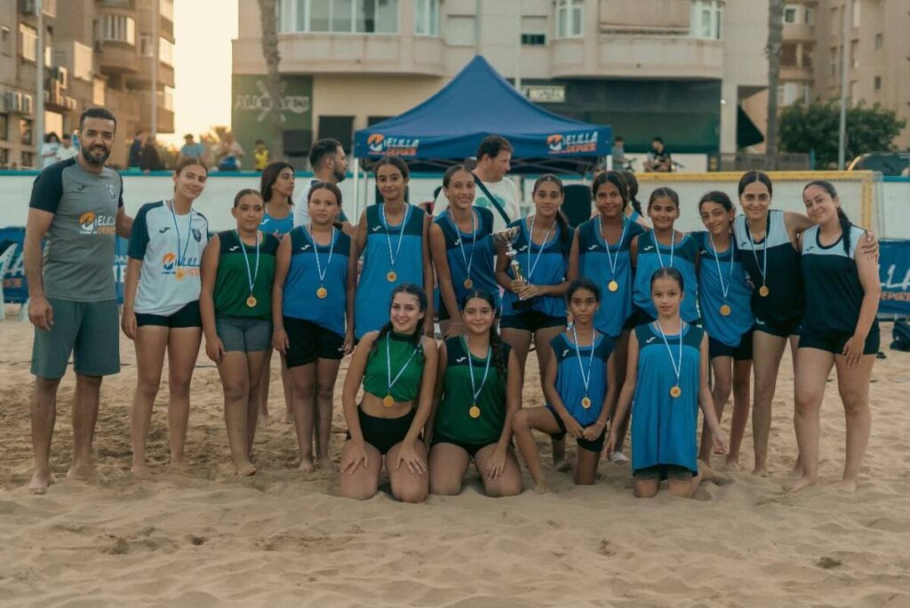 Youth girls' beach volleyball team posing with medals and a trophy on a sandy court, coach on the left.