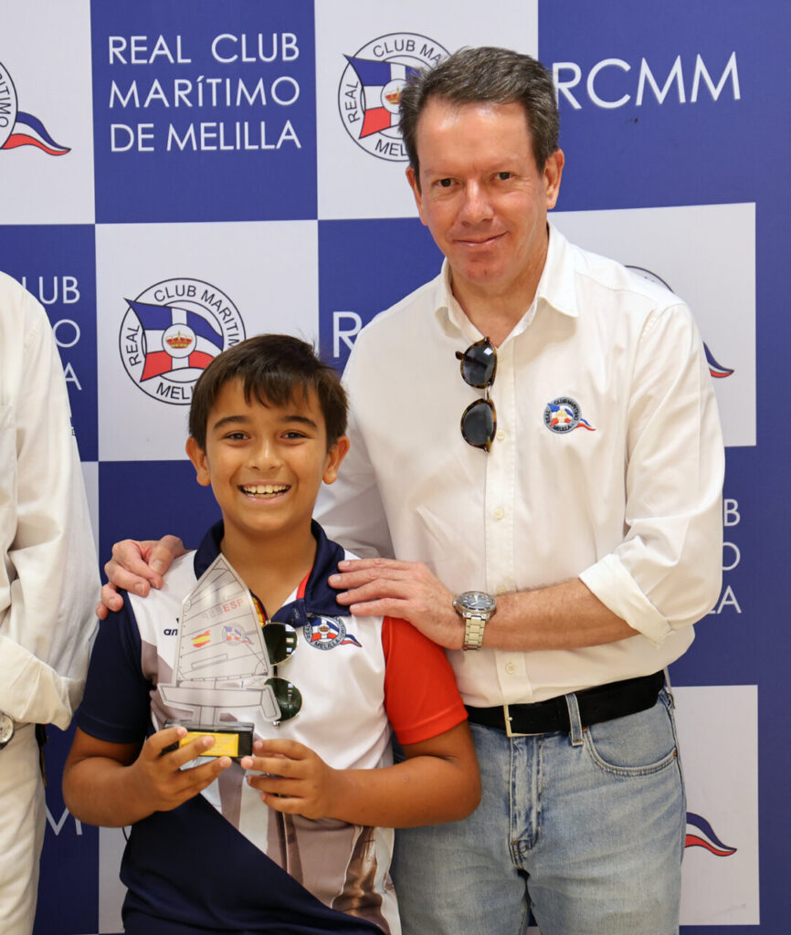 Boy smiling, holding a sailboat trophy at an award ceremony with a man in a white shirt beside him, backdrop shows RCMM logos.