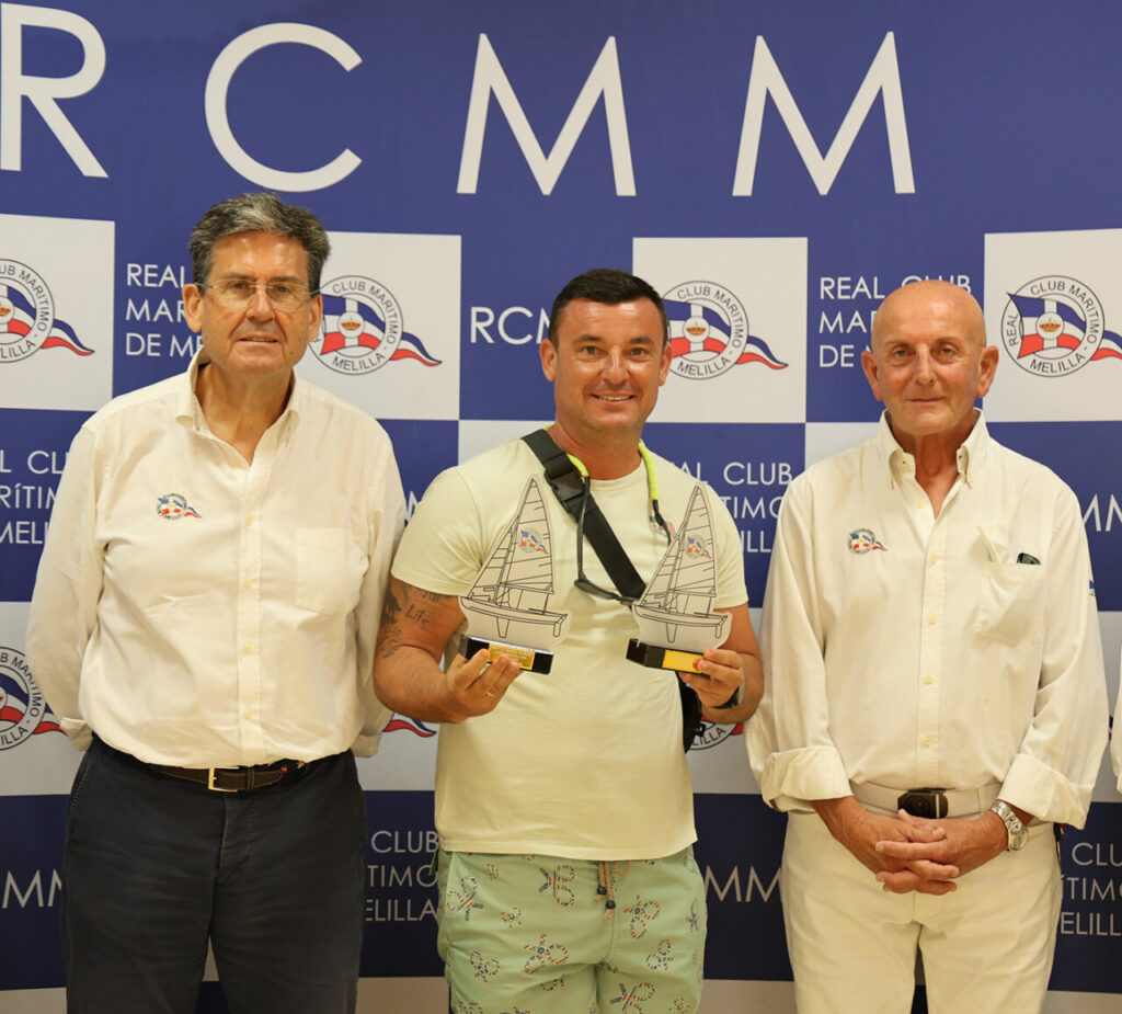 Three men pose for a photo at a maritime club event; the center man holds two sailboat trophies while flanked by two others in white shirts.
