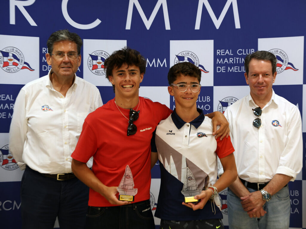 Two young sailors hold sailboat trophies while posing with two officials at a sailing club awards event, in front of a blue backdrop with club logos.