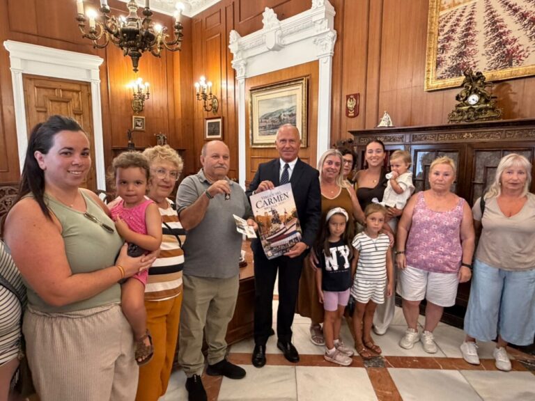 Group of adults and children posing together in a wood-paneled room; a man in a suit at center holds a poster titled 'Carmen'.