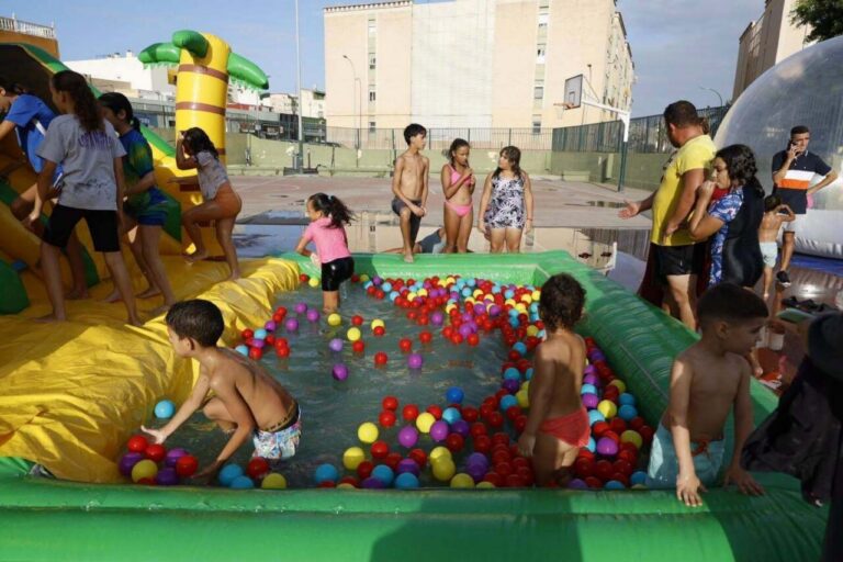 Kids play in a colorful inflatable pool filled with plastic balls at a sunny outdoor event, supervised by adults nearby.