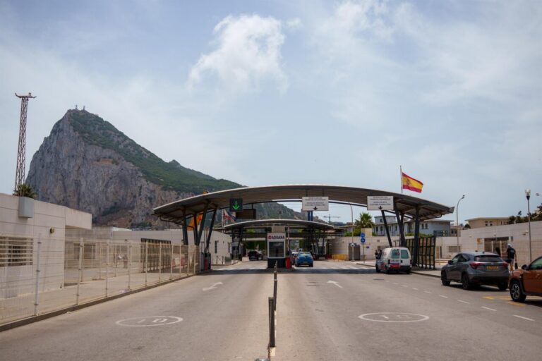 Border control checkpoint with a curved canopy over multiple lanes, vehicles waiting to be processed, and a Spanish flag in the background under a blue sky; rocky hill visible to the left.