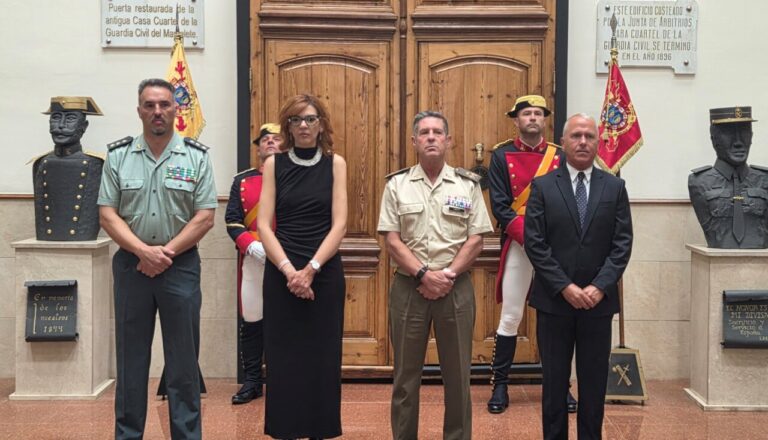 Four adults pose formally in a ceremonial hall in front of a large wooden door, flanked by guard statues and plaques on the walls in a dignified setting.