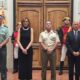 Four adults pose formally in a ceremonial hall in front of a large wooden door, flanked by guard statues and plaques on the walls in a dignified setting.