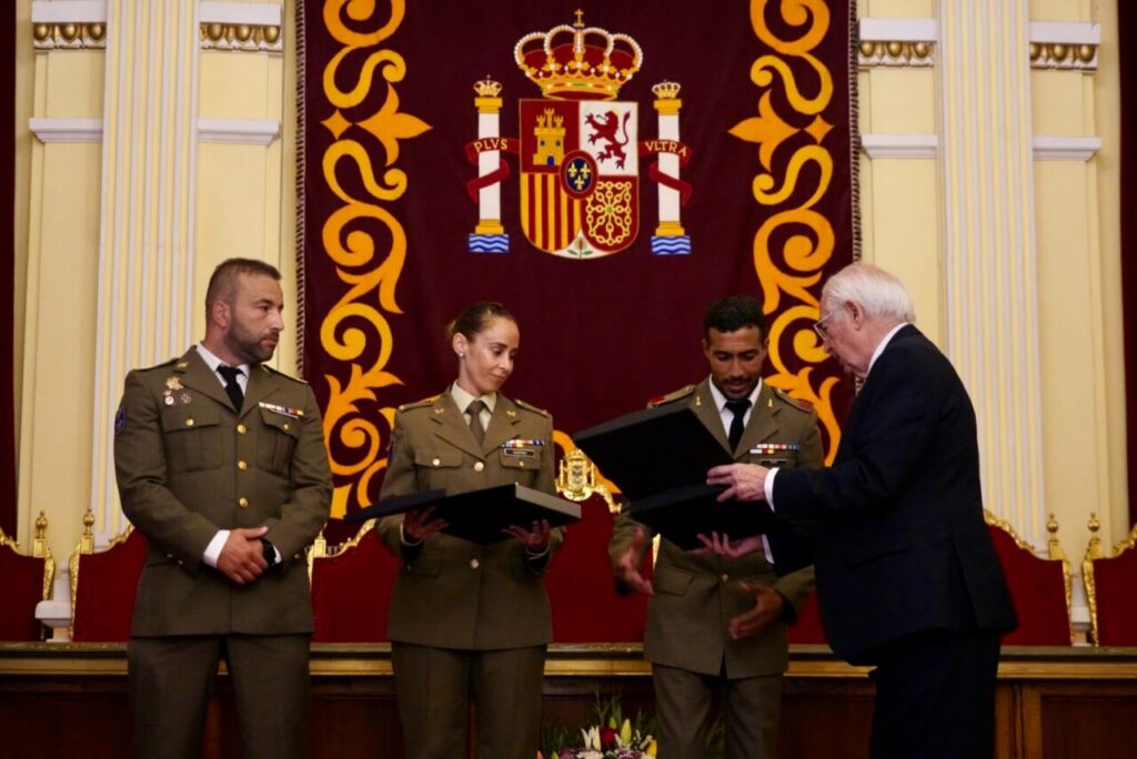 Military officers in uniform on a stage receive awards from a suited man, with a banner featuring a coat of arms behind them.