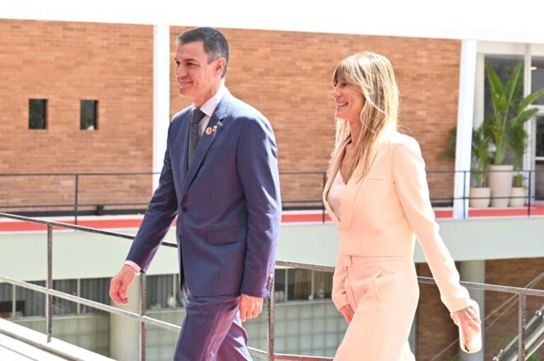 Two professionals walking outdoors, a man in a blue suit and a woman in a pale pink suit share a smile as they walk along a railing.
