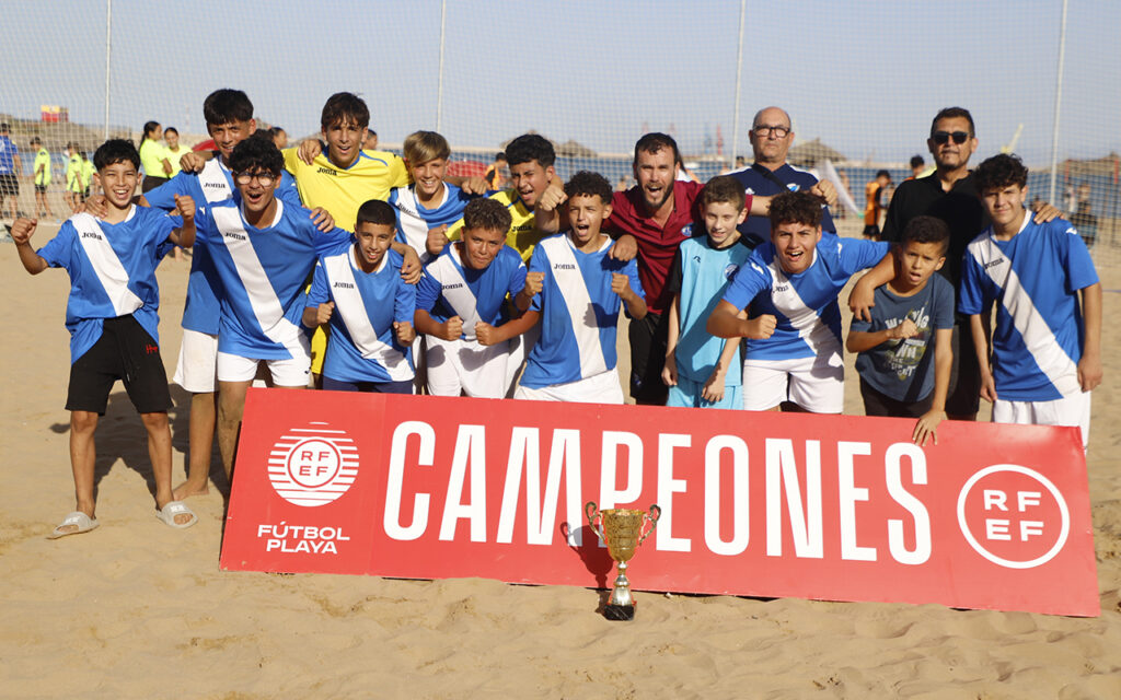 Youth beach soccer team posing with trophy behind a red 'CAMPEONES' banner, celebrating victory.