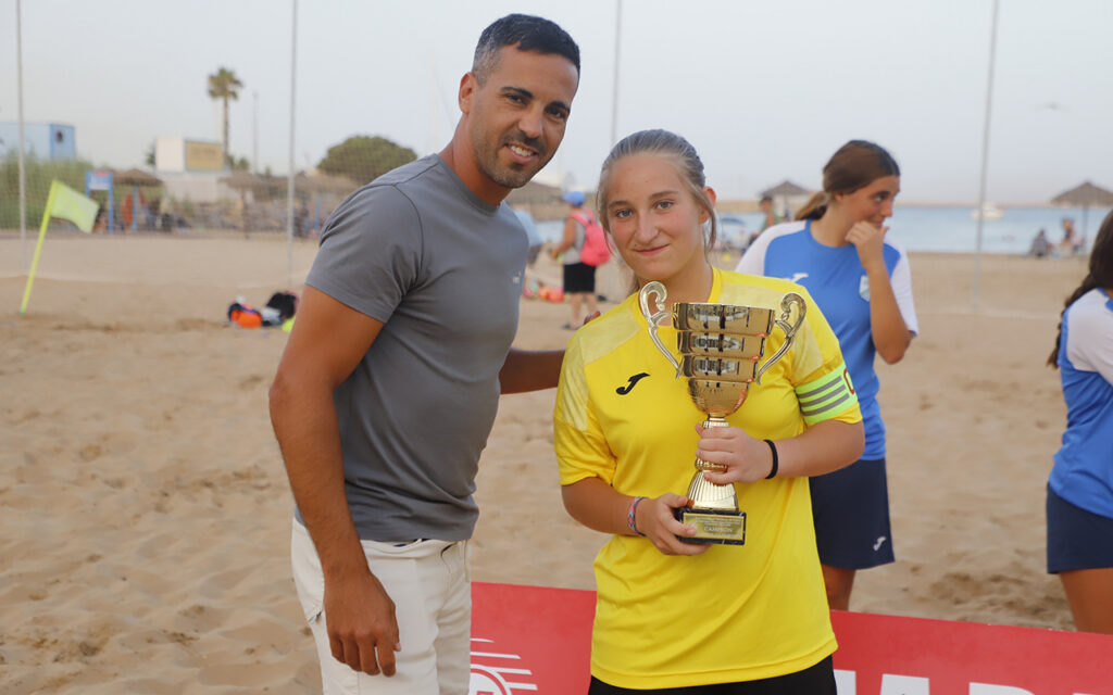 Man in a gray shirt stands beside a smiling girl in a yellow jersey, both on a sandy beach with a trophy in the girl's hands.