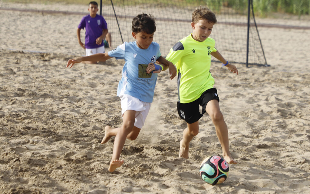 Two young boys play soccer on a sandy field, chasing a colorful ball as a teammate watches in the background.
