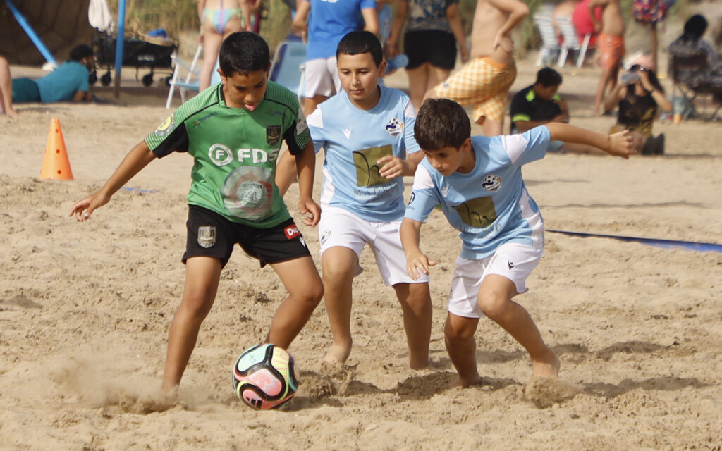 Three boys play beach soccer on sand, chasing a colorful ball with spectators in the background.