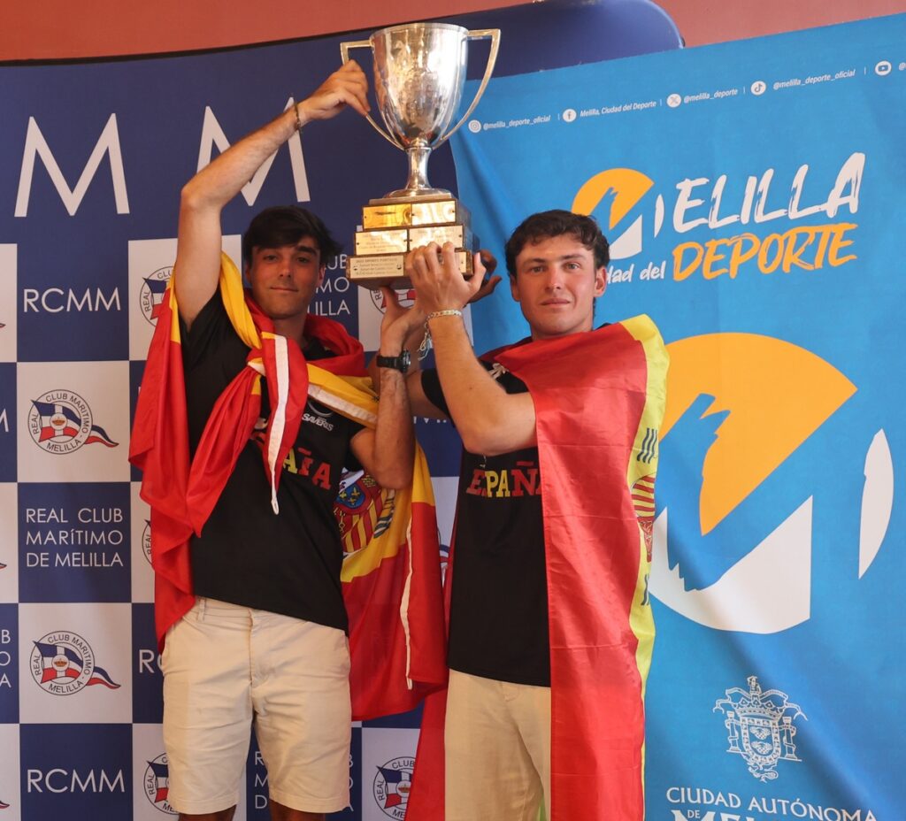 Two young men celebrate, lifting a large trophy while draped in Spanish flags at a sports event backdrop in Melilla.