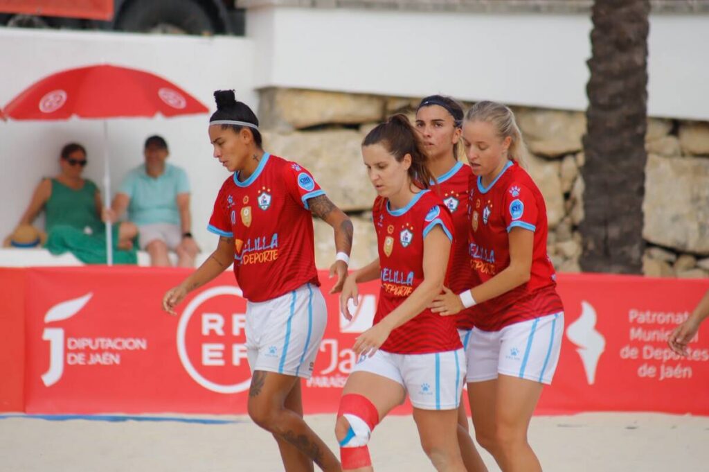 Four female beach soccer players in red jerseys with blue trim walk on a sandy court, spectators behind a red advertising barrier.