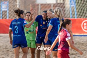 Group of women beach soccer players in blue celebrate together on a sandy field.