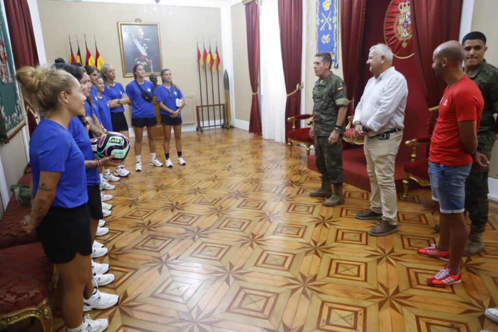 Women’s sports team in blue jerseys stands in a line, facing uniformed officials in a ceremonial room with flags and portraits in the background.