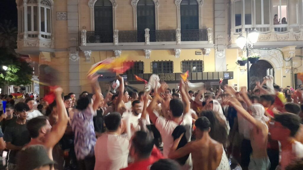 Crowd of people celebrating in a street at night, waving red‑yellow flags in front of a historic building with balconies.