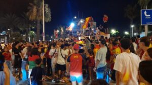 Nighttime street celebration with a dense crowd waving Spanish flags; people crowd around a float or stage with performers on top, some filming with phones.