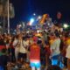 Nighttime street celebration with a dense crowd waving Spanish flags; people crowd around a float or stage with performers on top, some filming with phones.