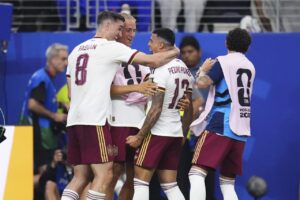 Group of Spain soccer players in white and maroon celebrate, embracing each other on the pitch after a goal.