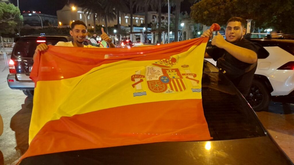 Two men hold a large Spanish flag over a car at night, smiling and giving thumbs up.