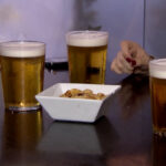 Tres vasos de cerveza sobre una mesa con un plato de snacks