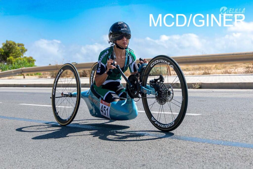 Handcycle race scene: athlete in green jersey and helmet pedals a blue handcycle on a sunny road, wearing number 451.