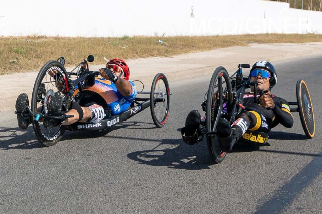 Two handcycle racing athletes lean into a turn on a road, wearing helmets; left rider in blue/orange kit, right rider in black/yellow with blue sunglasses.