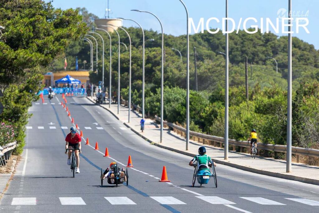 Cyclists ride along a closed road lined with orange cones during a sunny biking event; a handcycle appears in the foreground under street lamps.