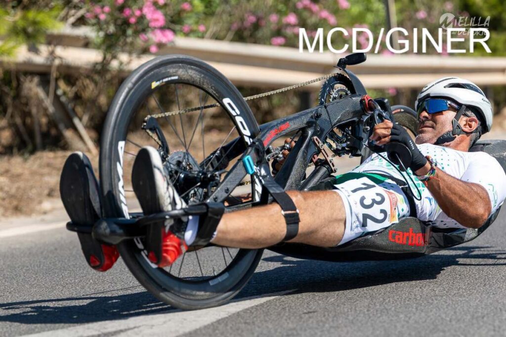 Paracyclist in a carbon handcycle racing on a road, wearing a white kit and helmet. (number 132 visible)