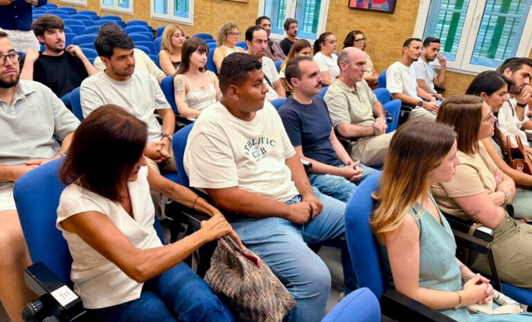 Group of adults seated in blue chairs listening to a talk in a classroom or lecture hall.