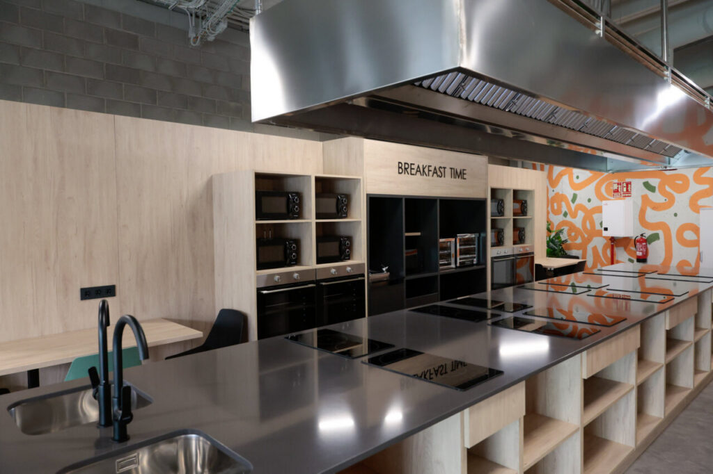 Industrial-style kitchen showroom with a long stainless steel island, built-in ovens and microwaves, and a 'BREAKFAST TIME' sign above the storage units.