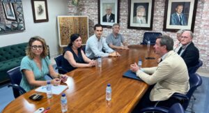 Group of seven adults seated around a large wooden conference table in a meeting room, with water bottles and papers on the table and framed portraits on a brick wall behind them.