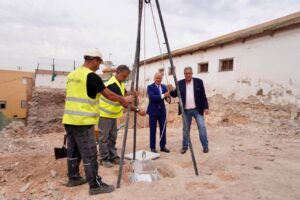 Group of workers and two suited men stand around a tripod drilling rig at a construction site, preparing to drill.