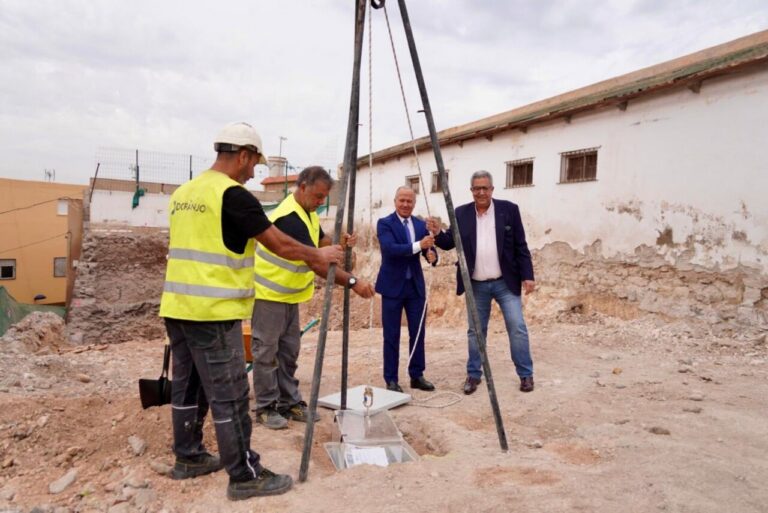Group of workers and two suited men stand around a tripod drilling rig at a construction site, preparing to drill.