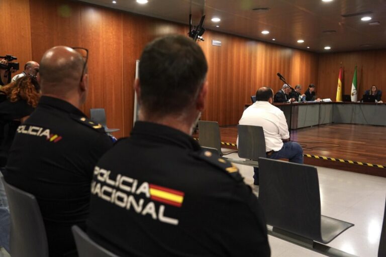 Panel of officials seated at a long table in a wood-paneled room; police officers in foreground watch the meeting.