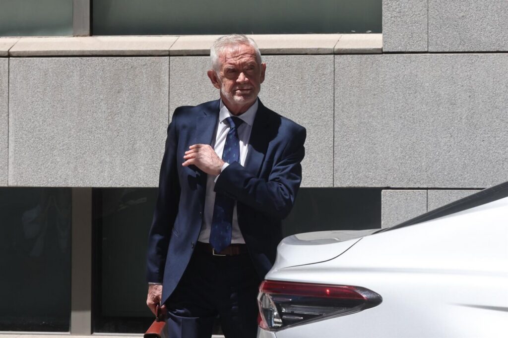 Older man in a navy suit adjusts his tie beside a white car outside a modern stone building.