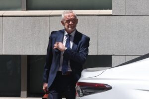 Older man in a navy suit adjusts his tie beside a white car outside a modern stone building.
