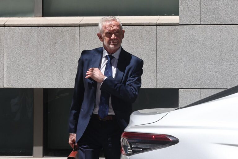Older man in a navy suit adjusts his tie beside a white car outside a modern stone building.