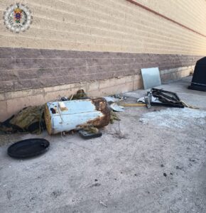 Abandoned outdoor scene with a rusted cylindrical tank and scattered debris along a beige brick wall; insulation material, wires, and a detached black lid nearby. A police emblem watermark appears in the upper left corner.