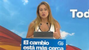 Woman speaking at a podium with two microphones and a tablet, in front of a blue backdrop that reads 'El cambio está más cerca' with a PP logo nearby.