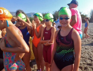 Group of young girls in swim caps and colorful swimsuits on a beach, awaiting a race start.