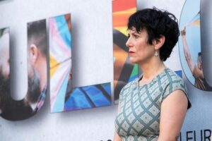 Side profile of a woman with short dark hair in a teal patterned dress, standing beside a colorful mural on a wall.