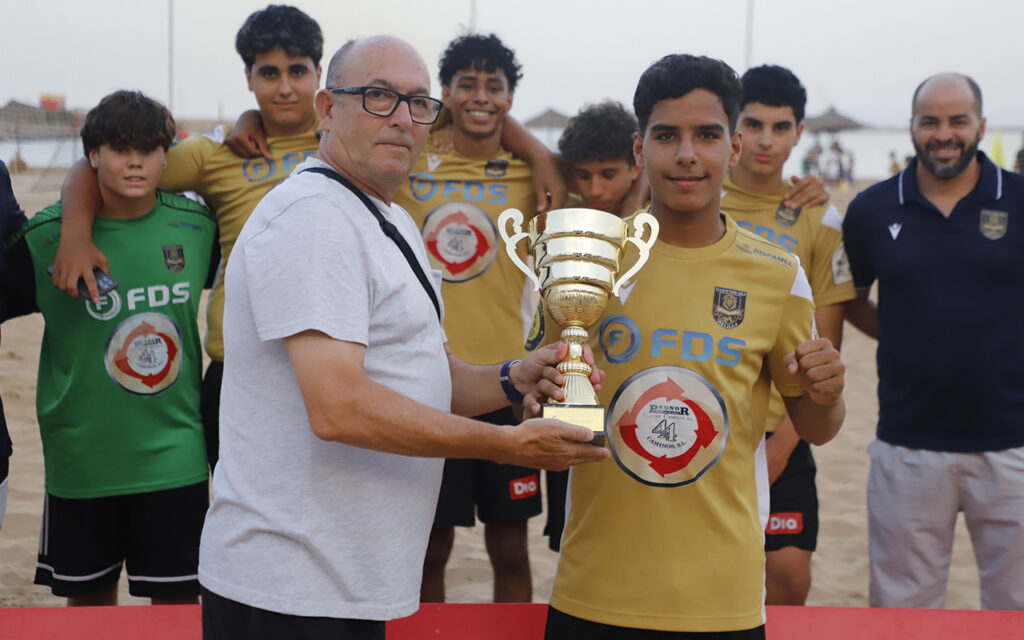 Coach hands a gold trophy to a young soccer player on a sandy beach as teammates celebrate in yellow jerseys behind them.