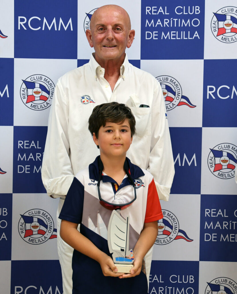 Two people pose in front of a blue-and-white checkered backdrop with logos, a tall man in a white shirt behind a smiling boy holding a sailboat trophy centerpiece