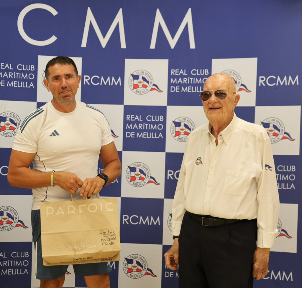 Two men pose for a photo in front of a blue RCMM backdrop; the younger man in a white athletic shirt holds a beige Parfois shopping bag while the older man wears sunglasses and a light shirt.
